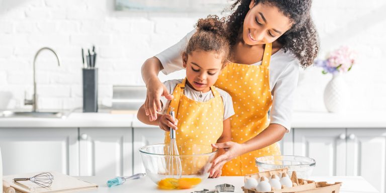 a young mother and daughter whisking an egg in a glass bowl, wearing matching yellow aprons in a white kitchen