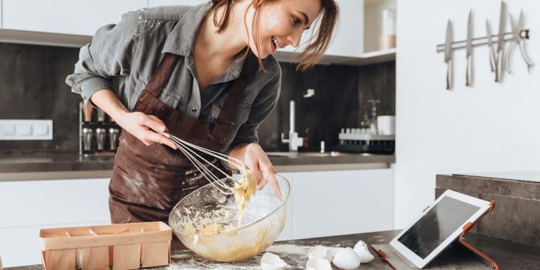 a woman looking at a tablet in the kitchen as she whisks a batter in a clear bowl