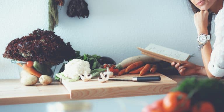 a cropped image of a woman reading a cookbook with vegetables and a cutting board on the table next to her