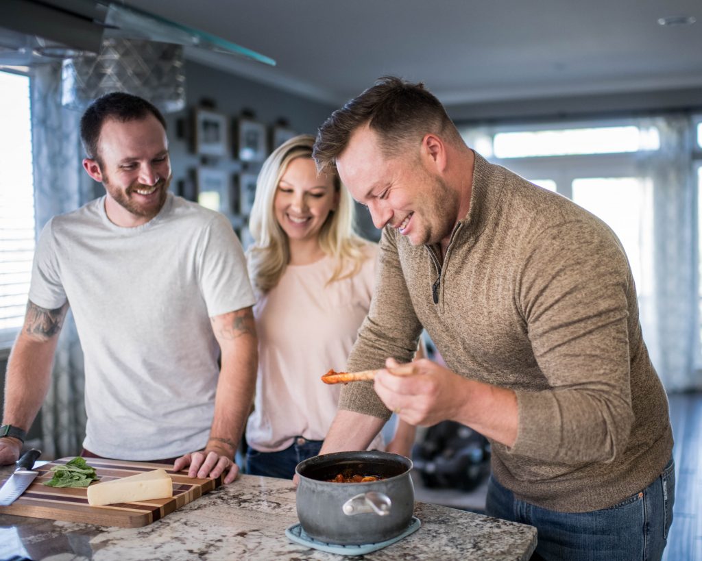 three young people gathered in a kitchen, smiling, as one taste tests a sauce