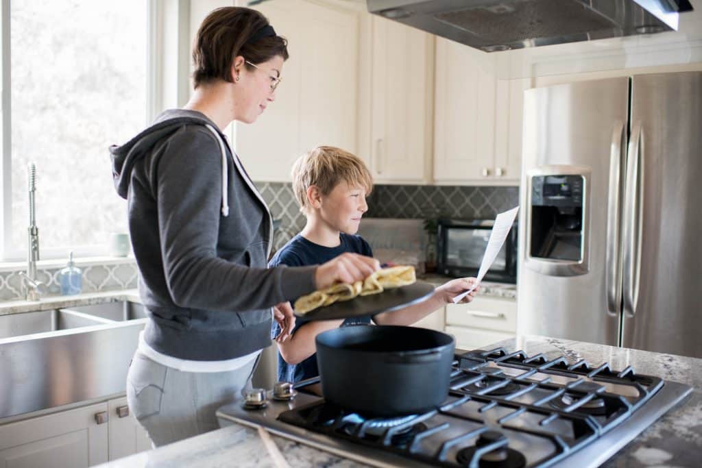 a woman holds open the lid of a pot, while son hold up recipe instructions on a piece of paper