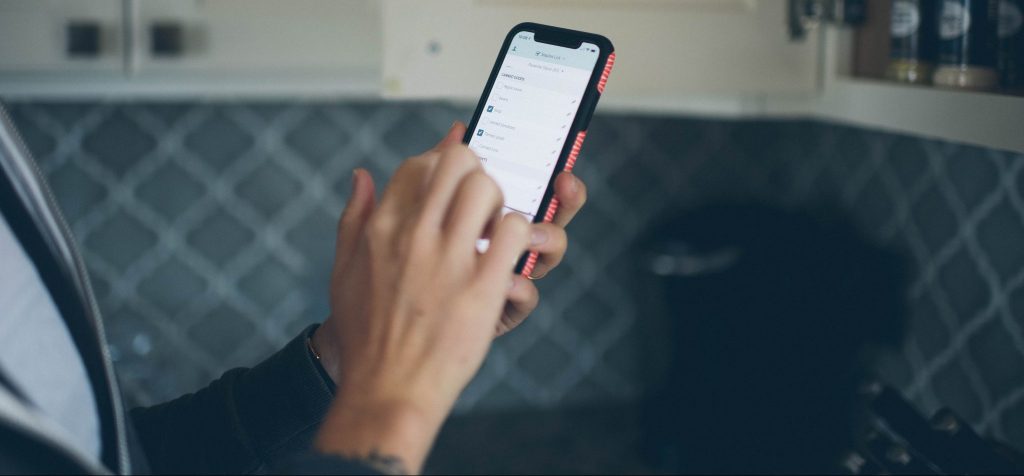 Cropped image of a hand holding a phone in a kitchen with a shopping list on screen