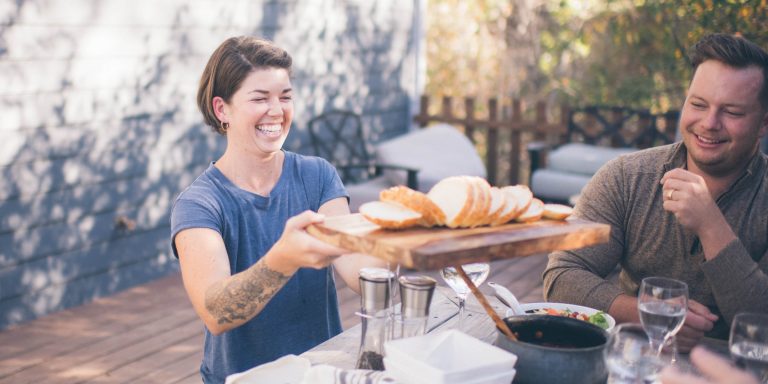 a woman in a blue shirt, smiling and passing a cutting board full of bread across the dinner table