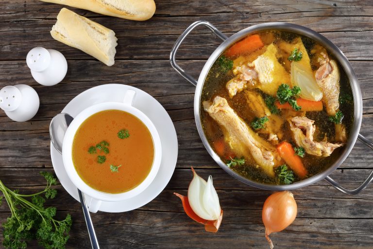 clear Chicken broth with pieces of rooster meat on bone and vegetables in a metal casserole and in a soup cup on dark wooden table, view from above, close-up