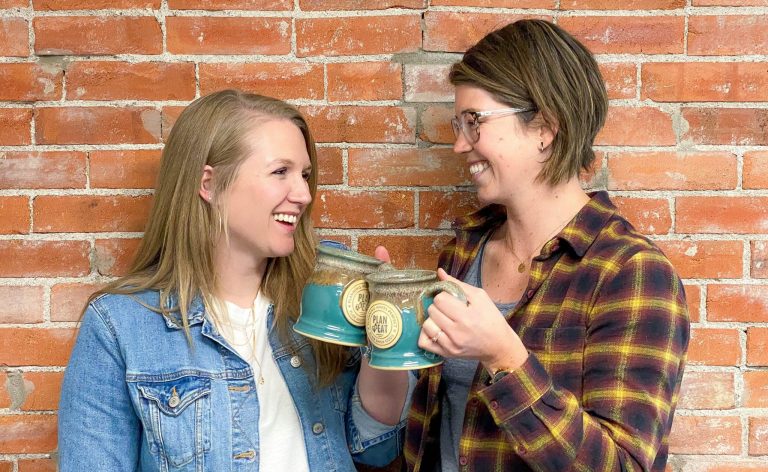 two young women cheersing blue mugs with a brick background