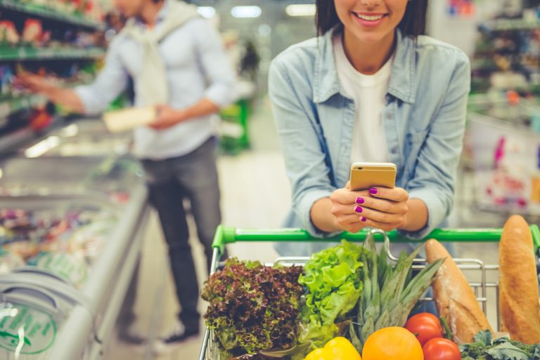 Couple in the supermarket. Cropped image of girl leaning on shopping cart, using a mobile phone and smiling, in the background her boyfriend is choosing food