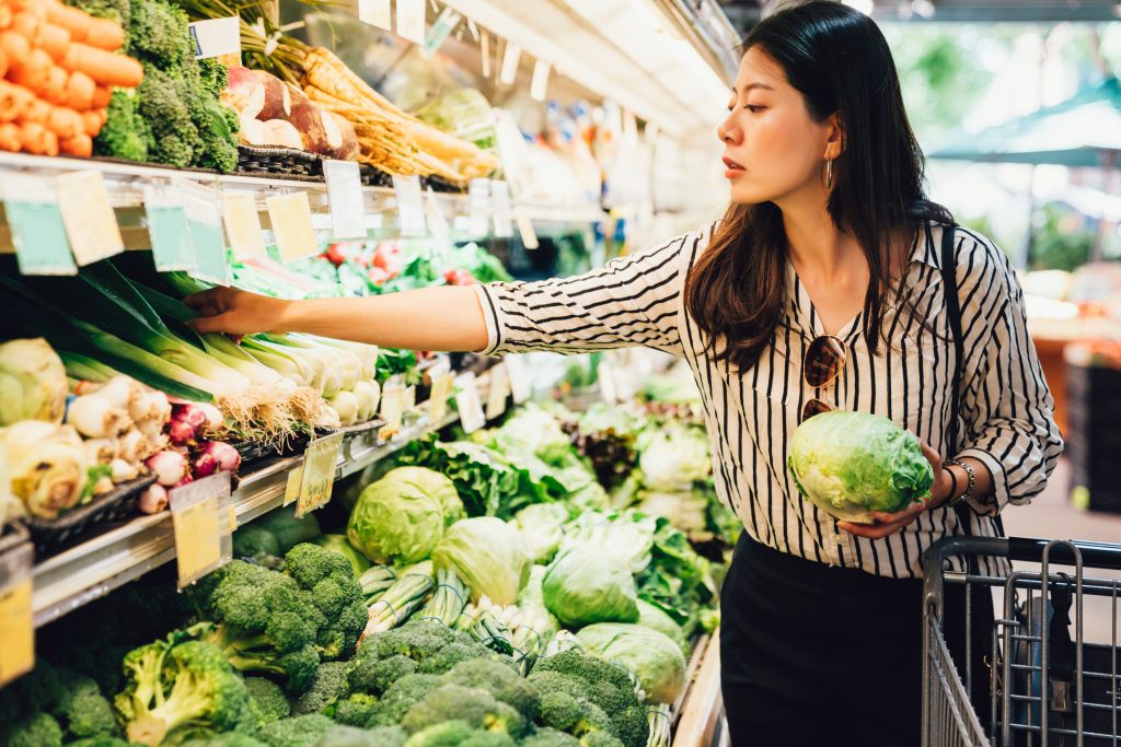 asian local woman buy vegetables and fruits in supermarket. young chinese lady holding green leaf vegetable and picking choosing green onion on cold open refrigerator. elegant female grocery shopping