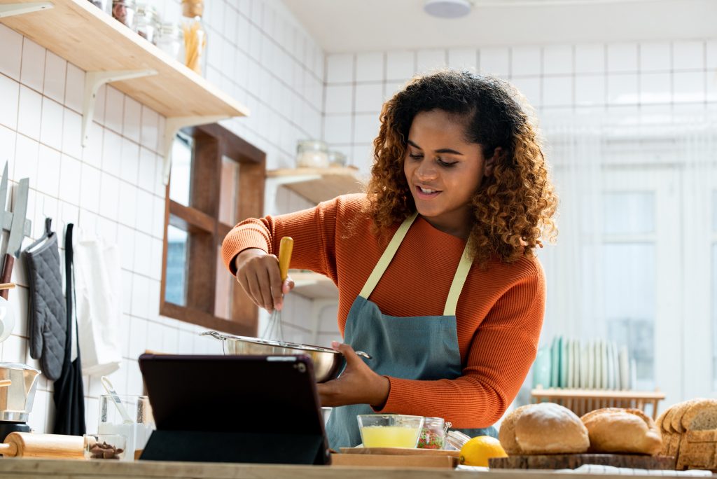 Young African American woman live streaming online with tablet computer while cooking in kitchen at home