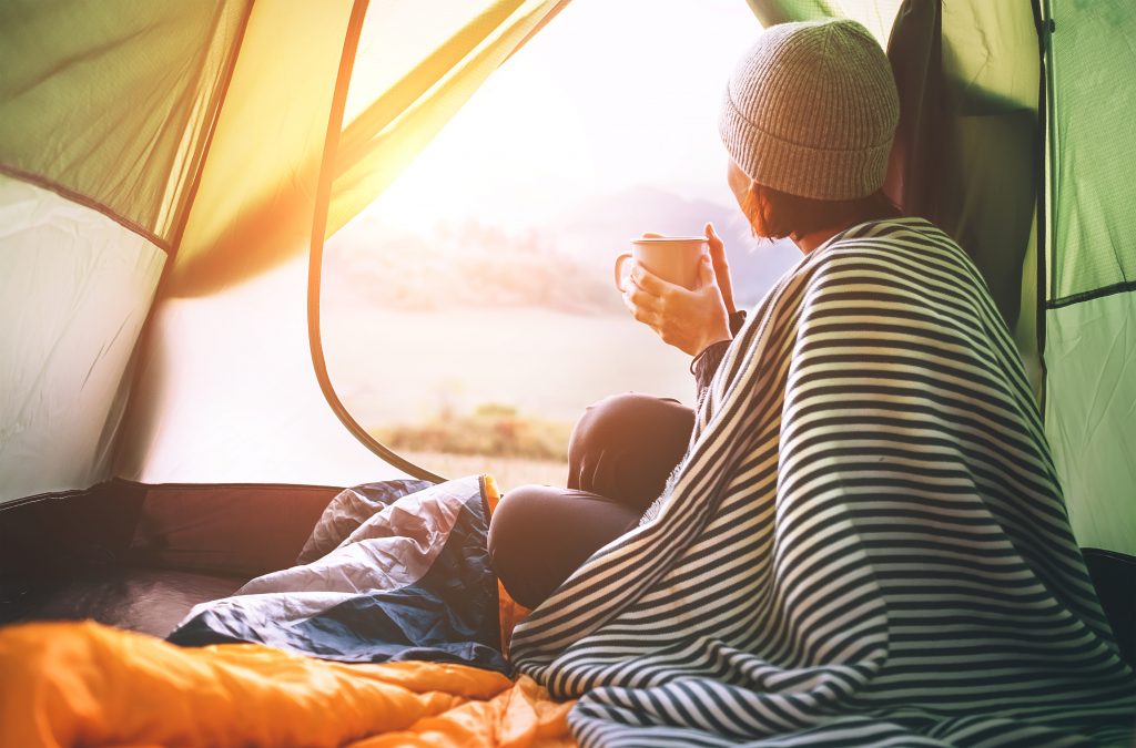 woman looks out the opening of a tent with a blanket on her shoulder and a hot drink in her hands