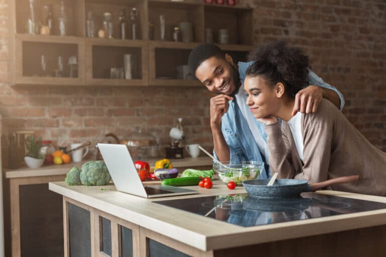 African-american couple cooking dinner with recipe on laptop in loft kitchen