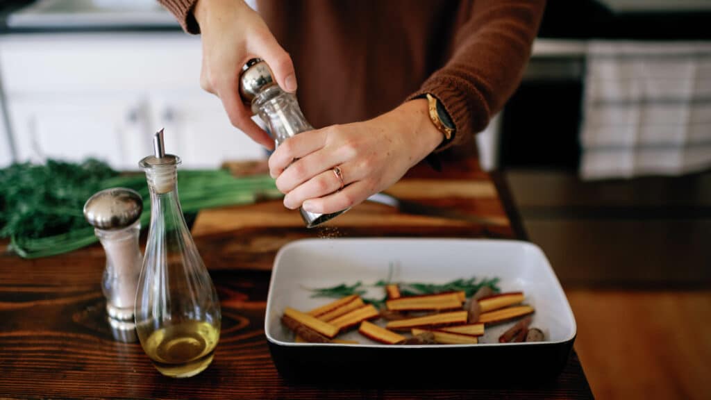 cropped image of woman adding fresh cracked pepper to sliced carrots in a baking dish