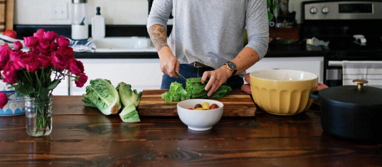 cropped image of woman chopping lettuce on a dark wood table with yellow bowl to the right and a vase of pink flowers to her left.